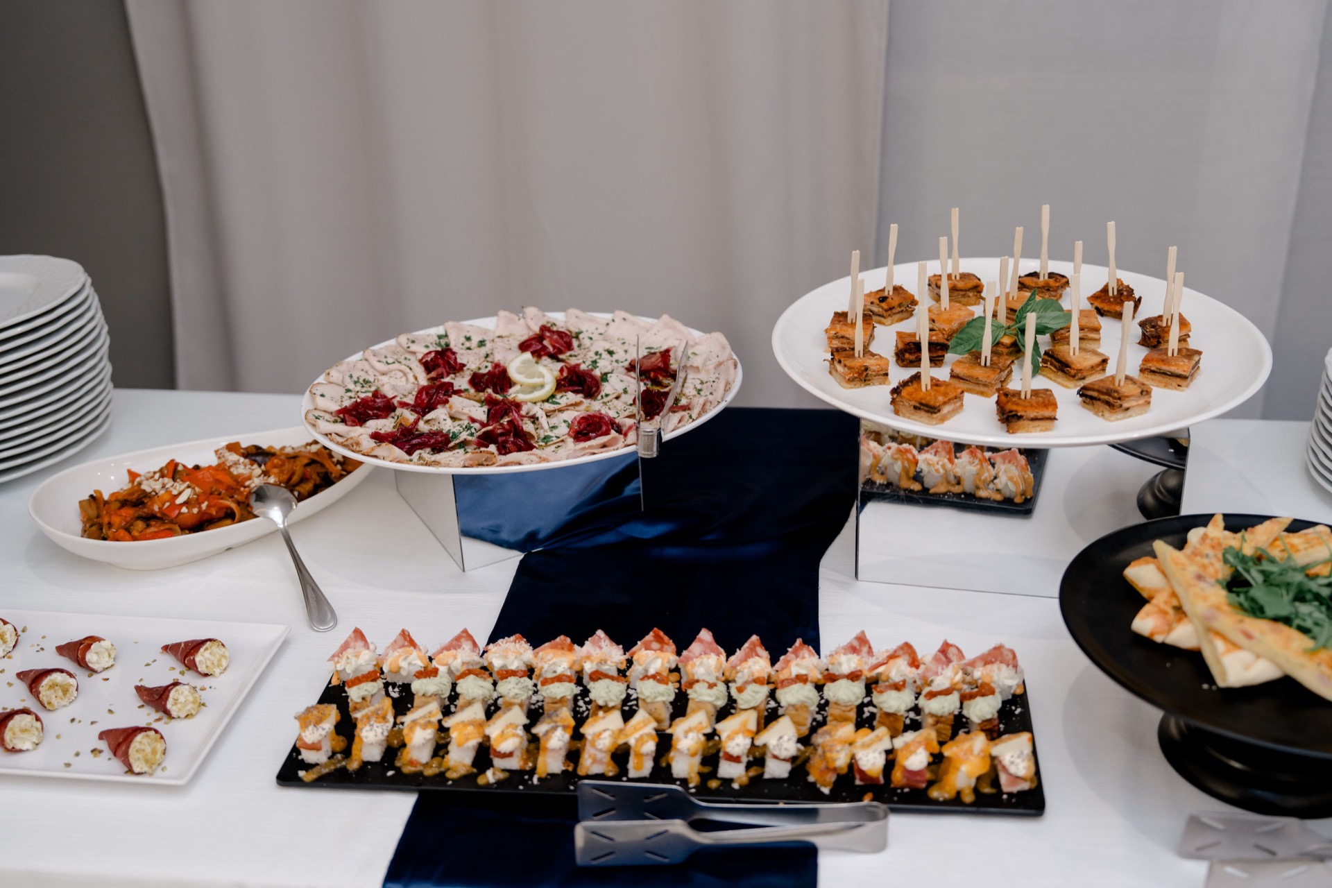 Tavolo buffet centrale con numerose portate fredde e calde disposte su alzate di vetro, piatti bianchi e vassoi neri. Si vedono finger food, salumi, formaggi, mini tramezzini e antipasti monoporzione in una presentazione ricca e ordinata da catering.