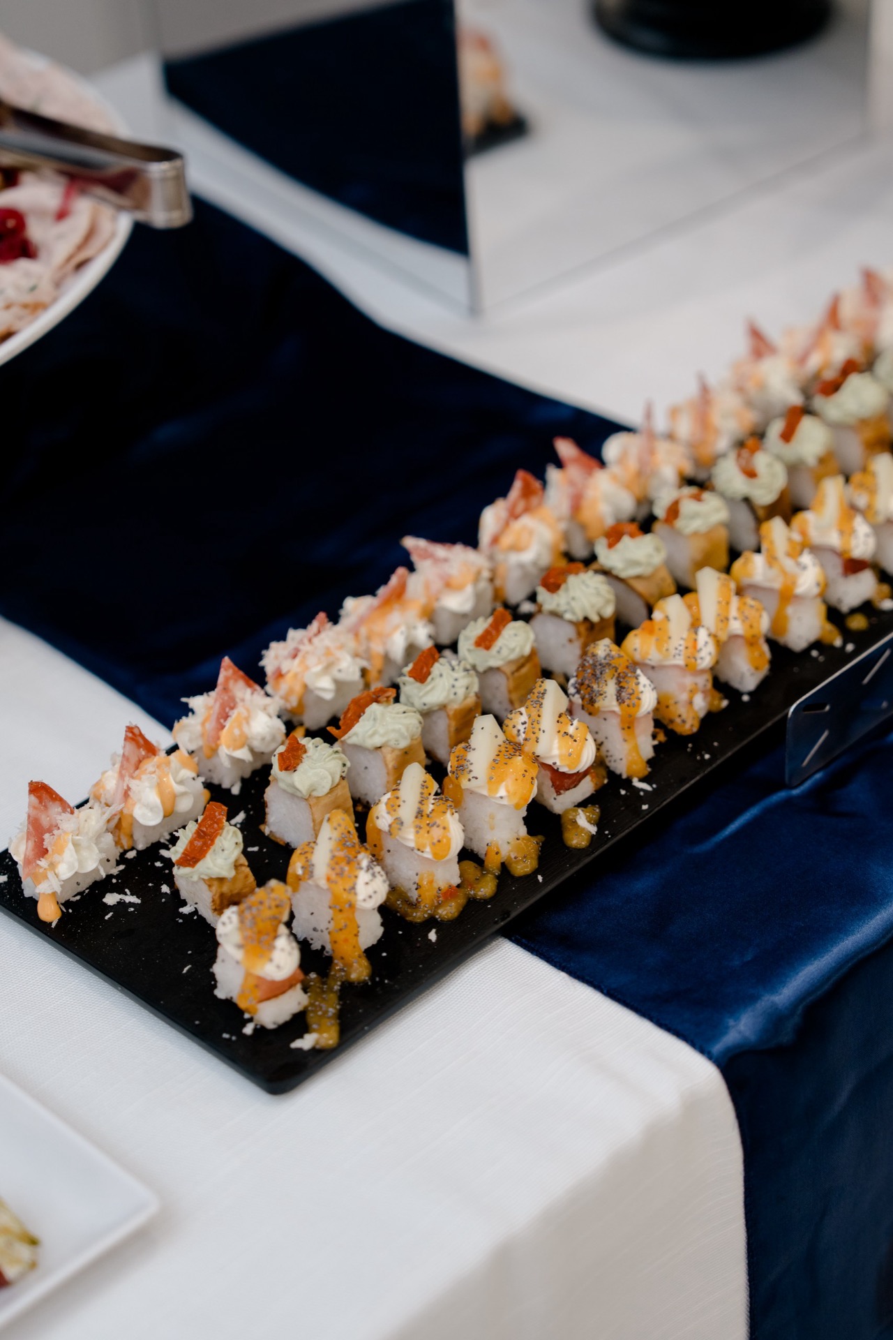 Vassoio nero con finger food tipo sushi o piccoli canapè disposti in file ordinate, decorati con salse chiare e arancioni. La composizione è molto ravvicinata e mostra una presentazione elegante da buffet serale.