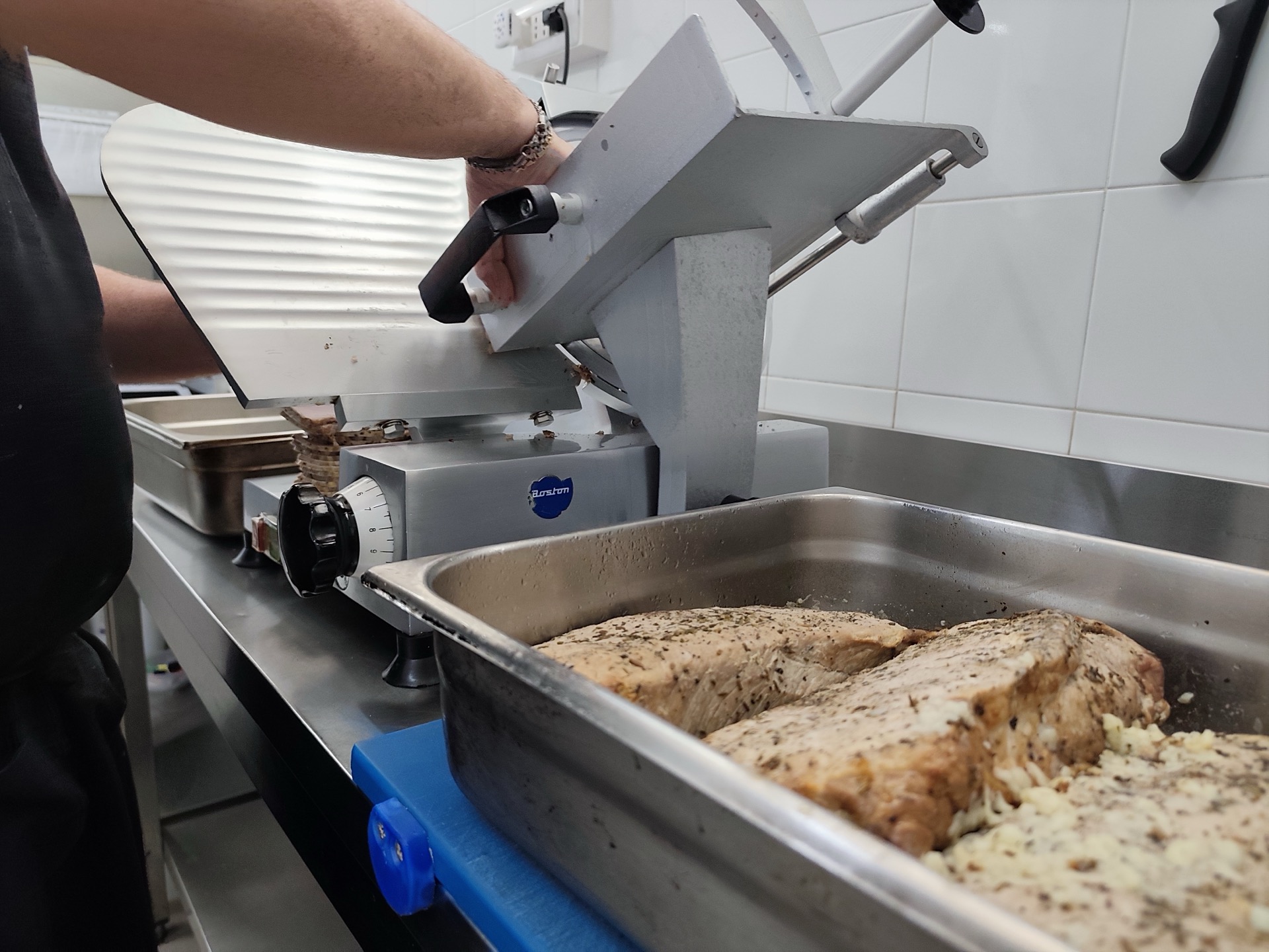 Preparazione in cucina con affettatrice professionale in uso e teglia di arrosto speziato in primo piano. La scena mostra una fase operativa del catering Viglianisi durante il taglio della carne prima del servizio.