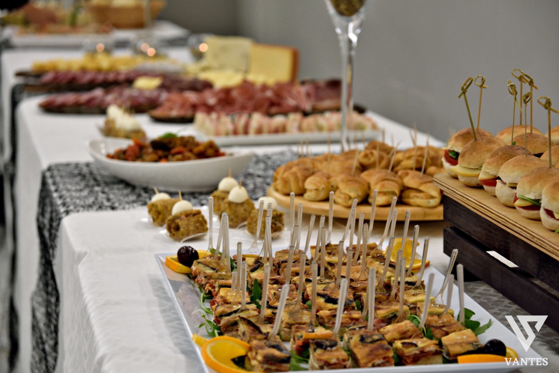 Apericena in sala con finger food, mini panini, rustici e vassoi caldi disposti su più tavoli di servizio. Sullo sfondo si vedono salumi, antipasti e altre portate da buffet, in un ambiente luminoso e ordinato per il catering.
