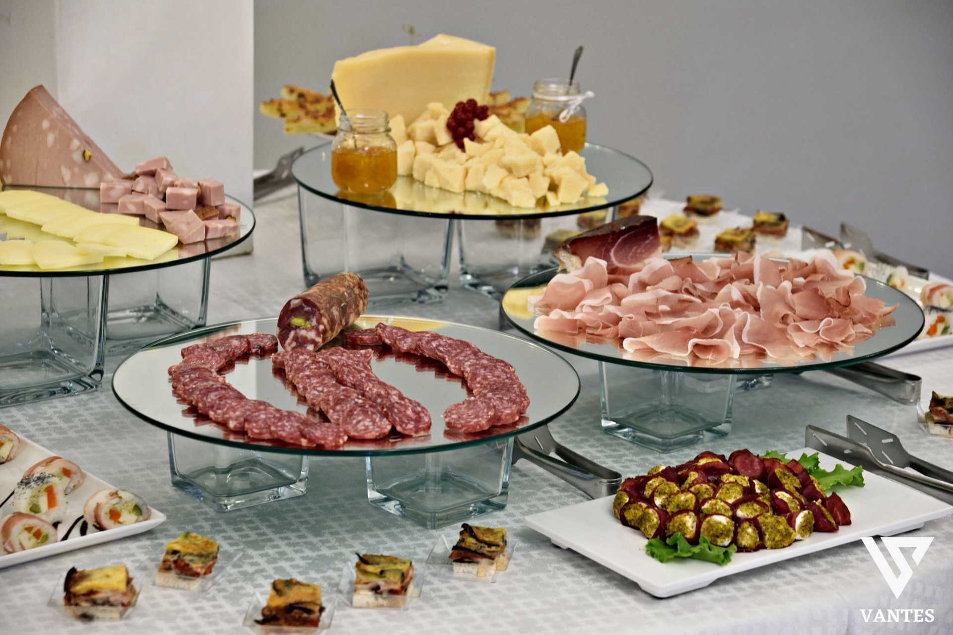 Buffet di catering con salumi, formaggi, finger food e piatti monoporzione esposti su alzate di vetro e vassoi bianchi. La tavola è ricca e ordinata, con presentazione elegante e piatti pronti al servizio.