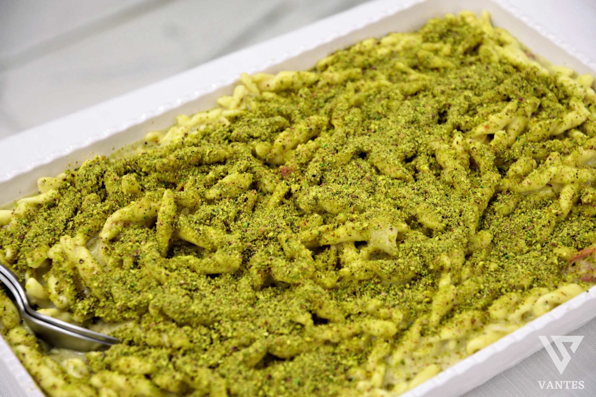 Vasca con pasta al pistacchio servita come primo piatto al buffet, ripresa molto da vicino e coperta da una granella verde uniforme. Il piatto è bianco e la preparazione mostra una presentazione semplice ma ricca di sapore.
