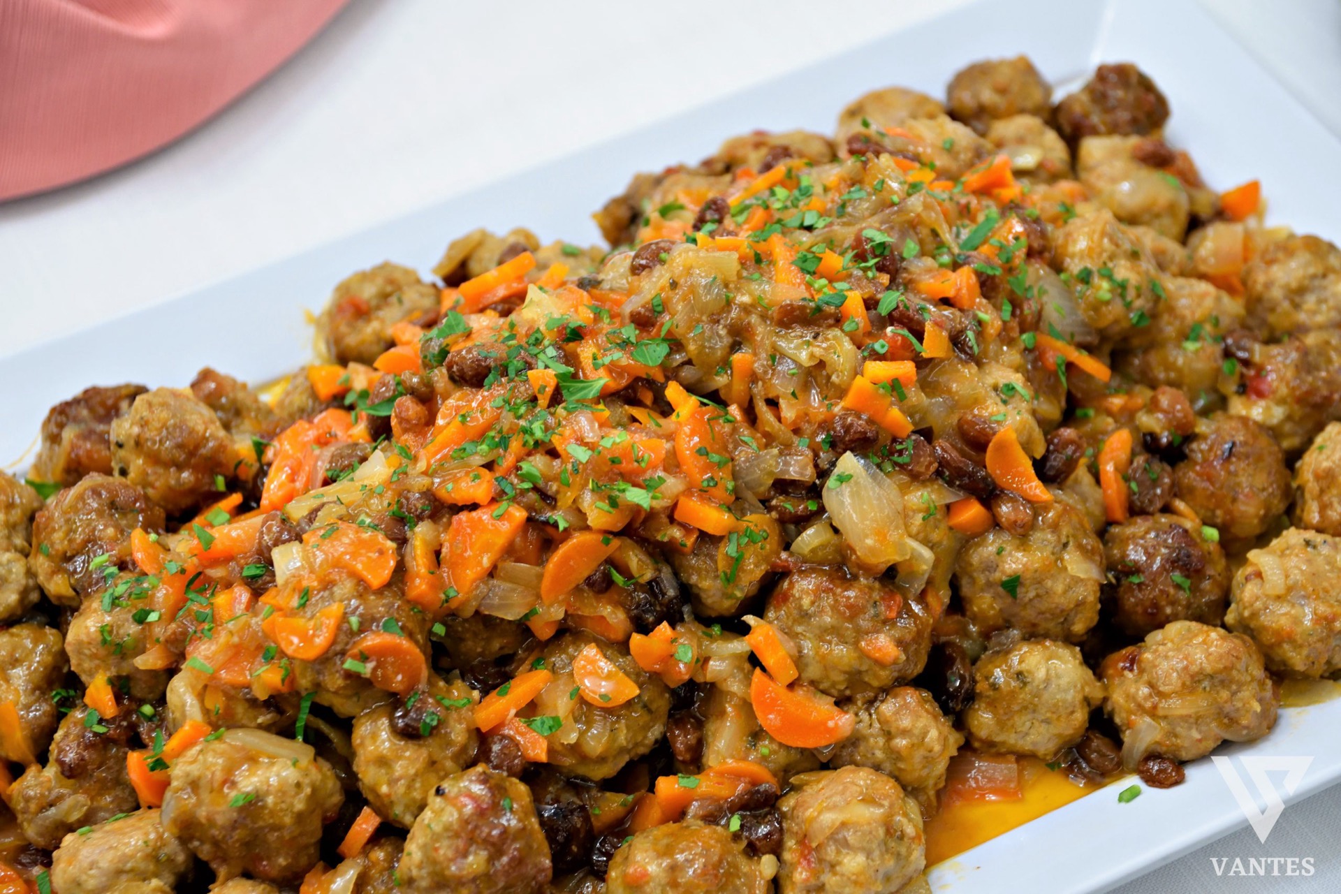 Secondo piatto al buffet con numerose polpette in umido condite con verdure, cipolle, uvetta e salsa, servite in un grande vassoio bianco. La preparazione è abbondante, colorata e molto ravvicinata.