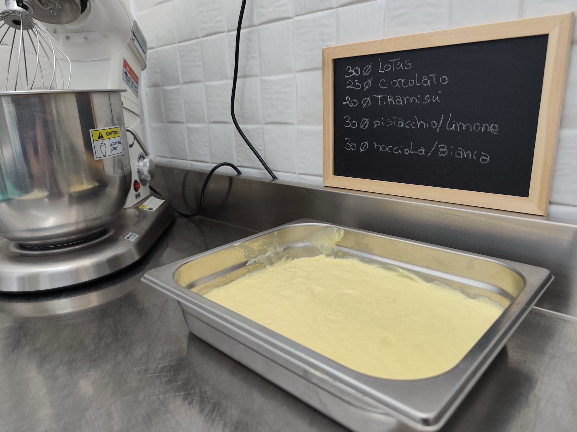 Teglia di crema pasticcera appena preparata su banco in acciaio, accanto a un'impastatrice professionale e a una lavagna con indicazioni di gusti e diametri delle torte. La foto documenta una fase di laboratorio della pasticceria di Viglianisi Catering.
