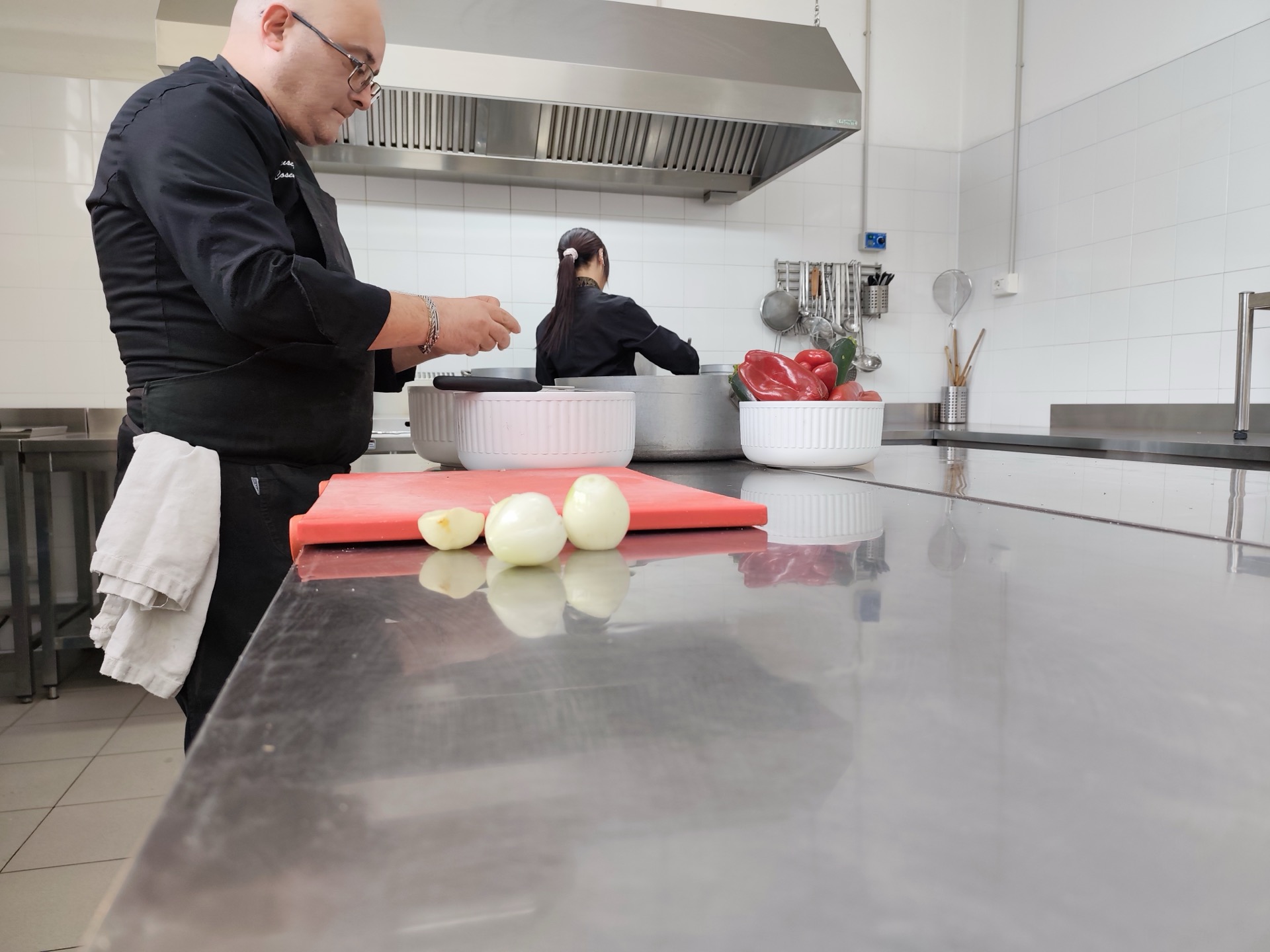 Cuochi al lavoro nella cucina professionale di Viglianisi Catering, tra pentole, banco inox e ingredienti pronti alla preparazione. In primo piano si vedono cipolle e peperoni, a sottolineare la fase operativa del servizio cucina.