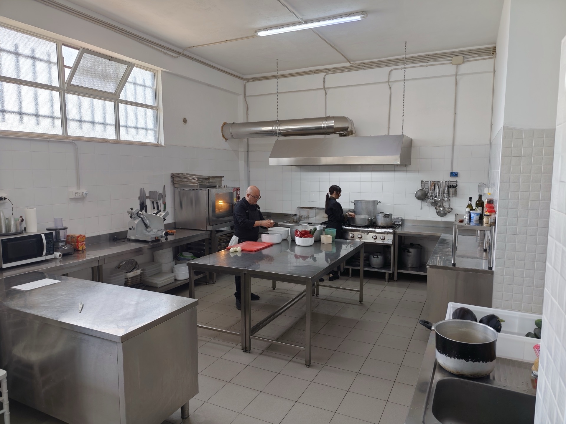 Vista ampia della cucina professionale di Viglianisi Catering, con cuochi al lavoro tra fornelli, banco inox, pentole e attrezzature. L'immagine racconta l'organizzazione del laboratorio in una fase di preparazione operativa.