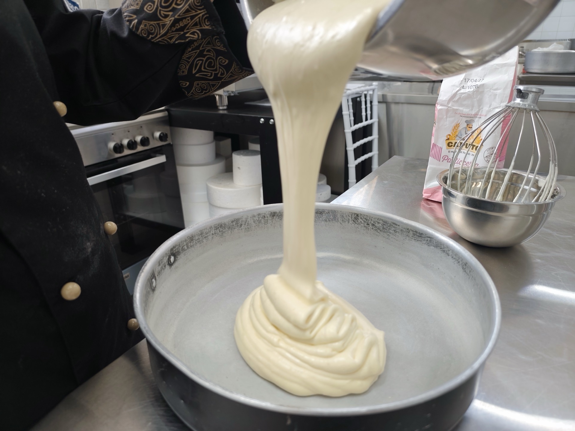 Impasto chiaro e denso versato in una tortiera durante una preparazione di pasticceria, con frusta, ciotola e farina sul banco di lavoro. La foto mostra una fase artigianale del laboratorio dolci di Viglianisi Catering.