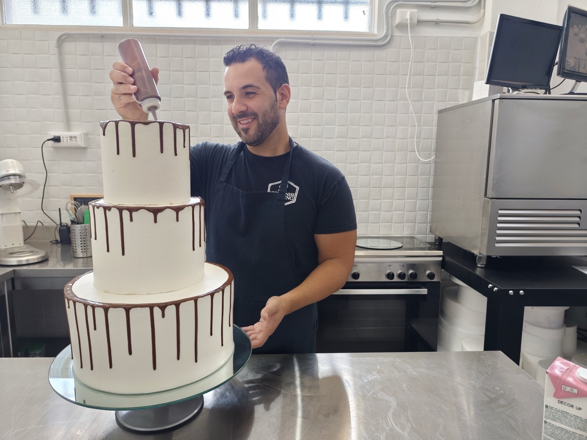 Pasticcere al lavoro su una drip cake bianca a tre piani, mentre applica la colatura di cioccolato lungo il bordo superiore. L'immagine valorizza la cura artigianale della pasticceria firmata Viglianisi Catering.