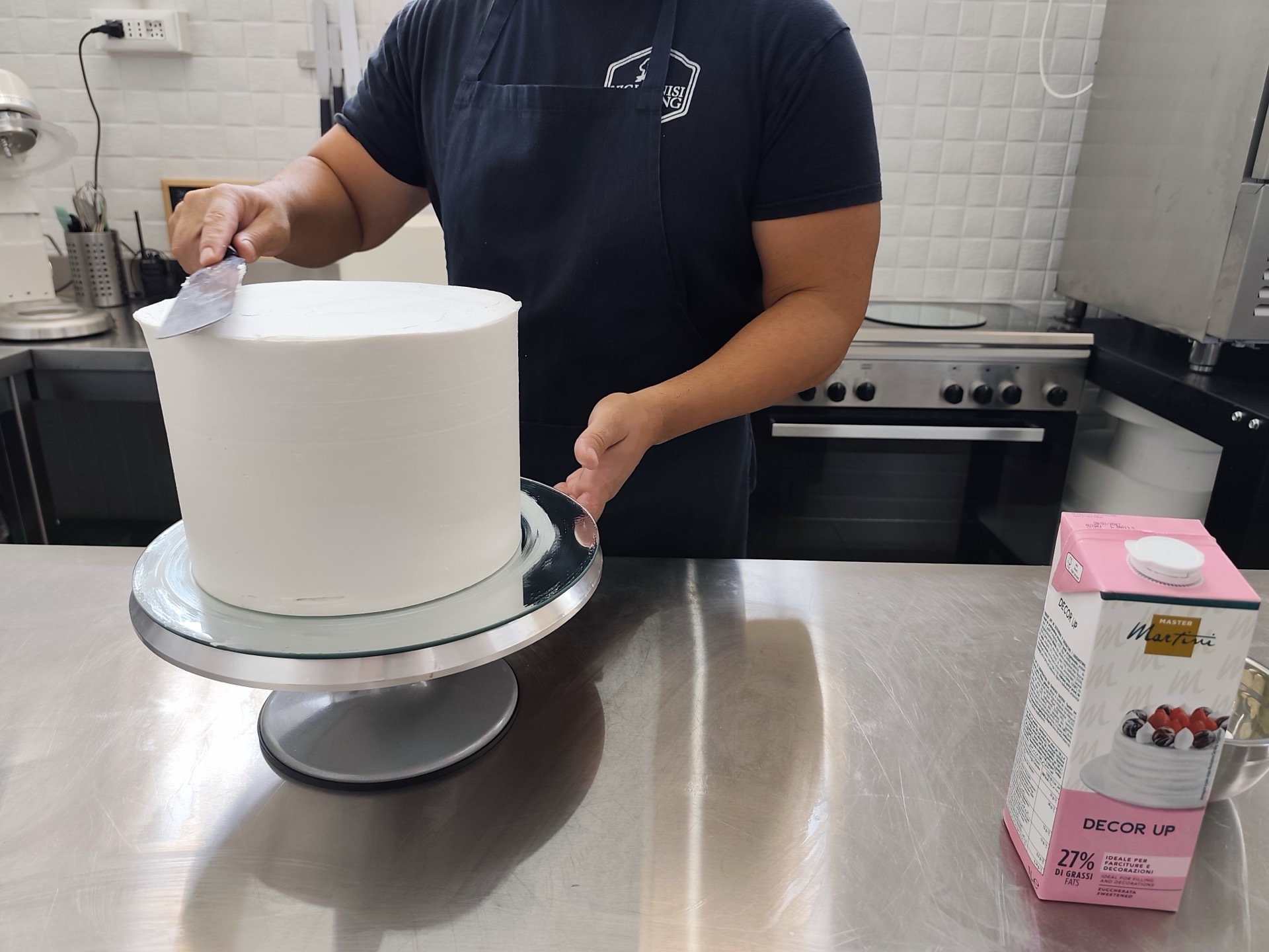 Pasticcere mentre rifinisce con spatola la copertura liscia di una grande torta bianca su alzata girevole. L'immagine racconta la fase di finitura della pasticceria artigianale di Viglianisi Catering.