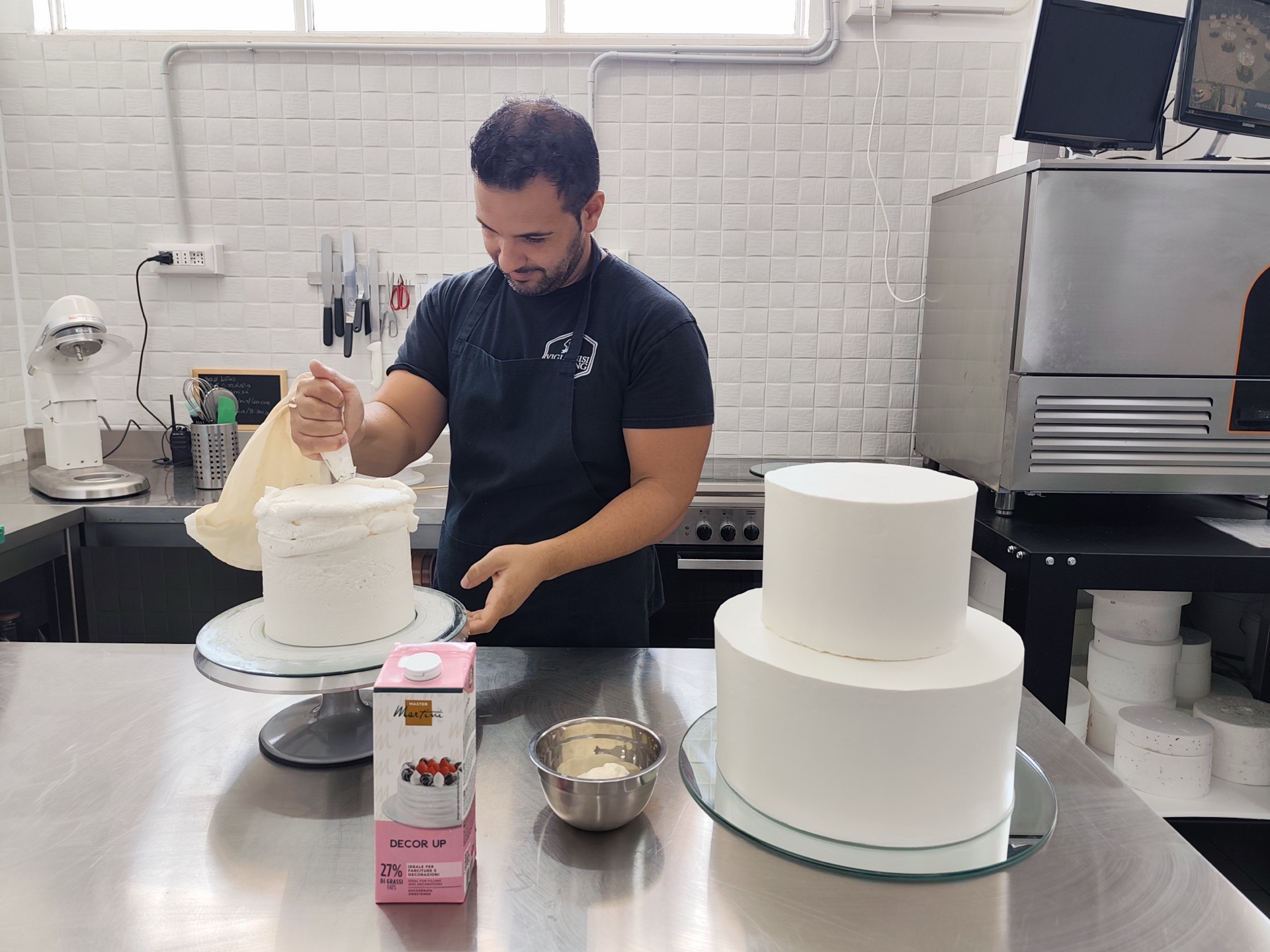 Pasticcere al lavoro con sac à poche su una torta in fase di montaggio, mentre sul banco accanto si vedono altre basi già rivestite di bianco. L'immagine documenta una fase avanzata della lavorazione artigianale in pasticceria.