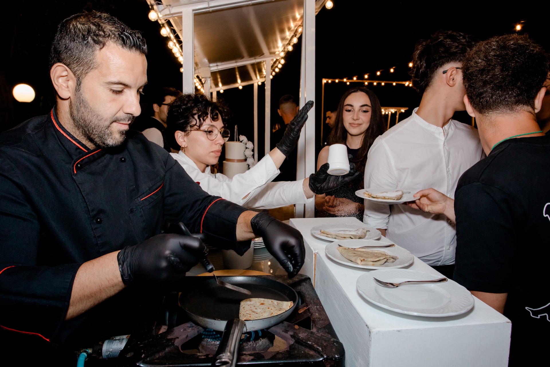 Chef che prepara crêpes al momento su una piastra, mentre ospiti e personale attendono il servizio lungo il banco. Sul tavolo ci sono piatti già pronti e l'atmosfera notturna è illuminata da catenarie calde all'esterno.