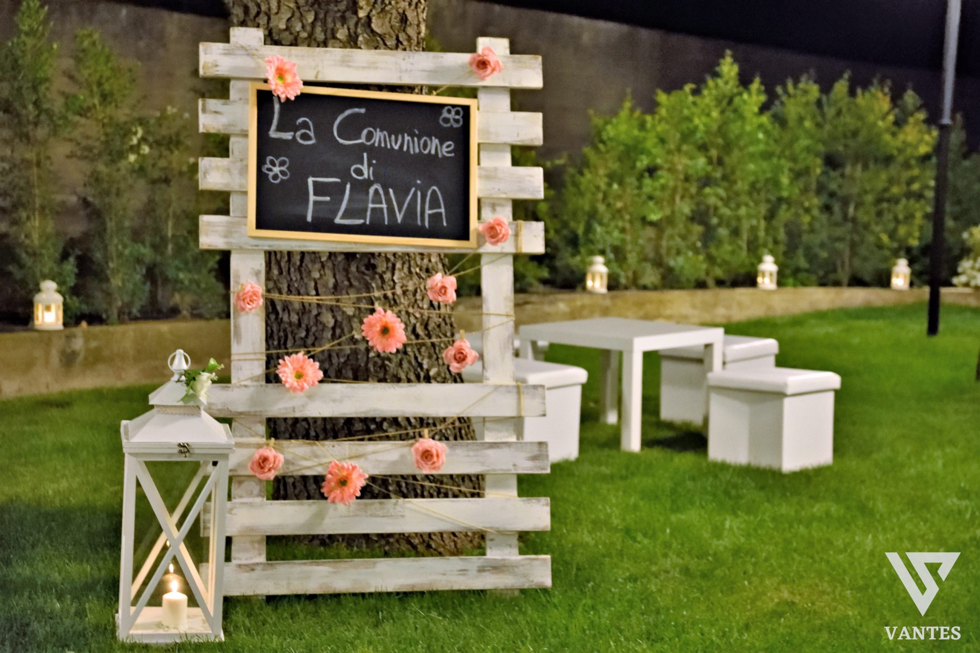 Scenografia per la comunione di Flavia allestita in giardino con pannello in legno chiaro, lavagna centrale decorata con fiori rosa e lanterna bianca a terra. Sullo sfondo ci sono tavolini e pouf bianchi su prato verde, con luci decorative lungo il perimetro.