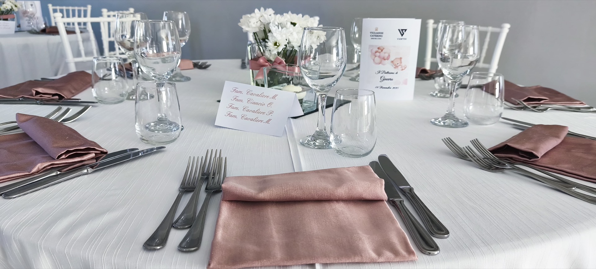 Dettaglio di un tavolo da battesimo apparecchiato con tovagliato bianco, tovaglioli rosa cipria, calici trasparenti e cartoncino centrale con il nome degli invitati. Al centro c'è anche un piccolo vaso con fiori bianchi, per una mise en place sobria e raffinata.