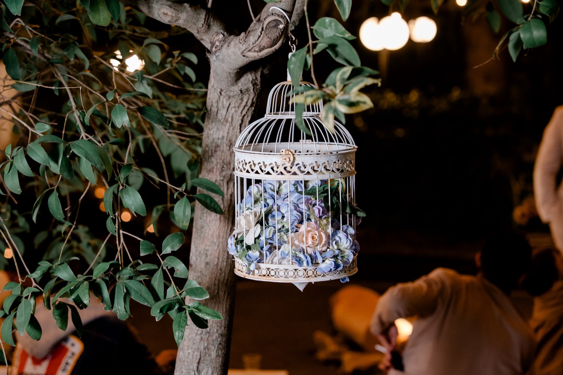 Gabbia decorativa bianca appesa a un albero nel giardino, riempita di fiori azzurri e rosa con fogliame verde. Lo sfondo notturno è illuminato da luci calde e la scena richiama un allestimento romantico per evento all'aperto.