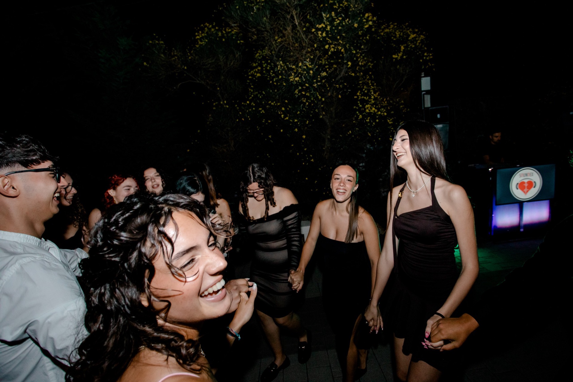 Gruppo di ragazzi che balla e ride durante una festa all'aperto di notte, con la postazione DJ visibile sullo sfondo. La scena è vivace e illuminata da luce flash e da piccoli punti luce caldi tra gli alberi.