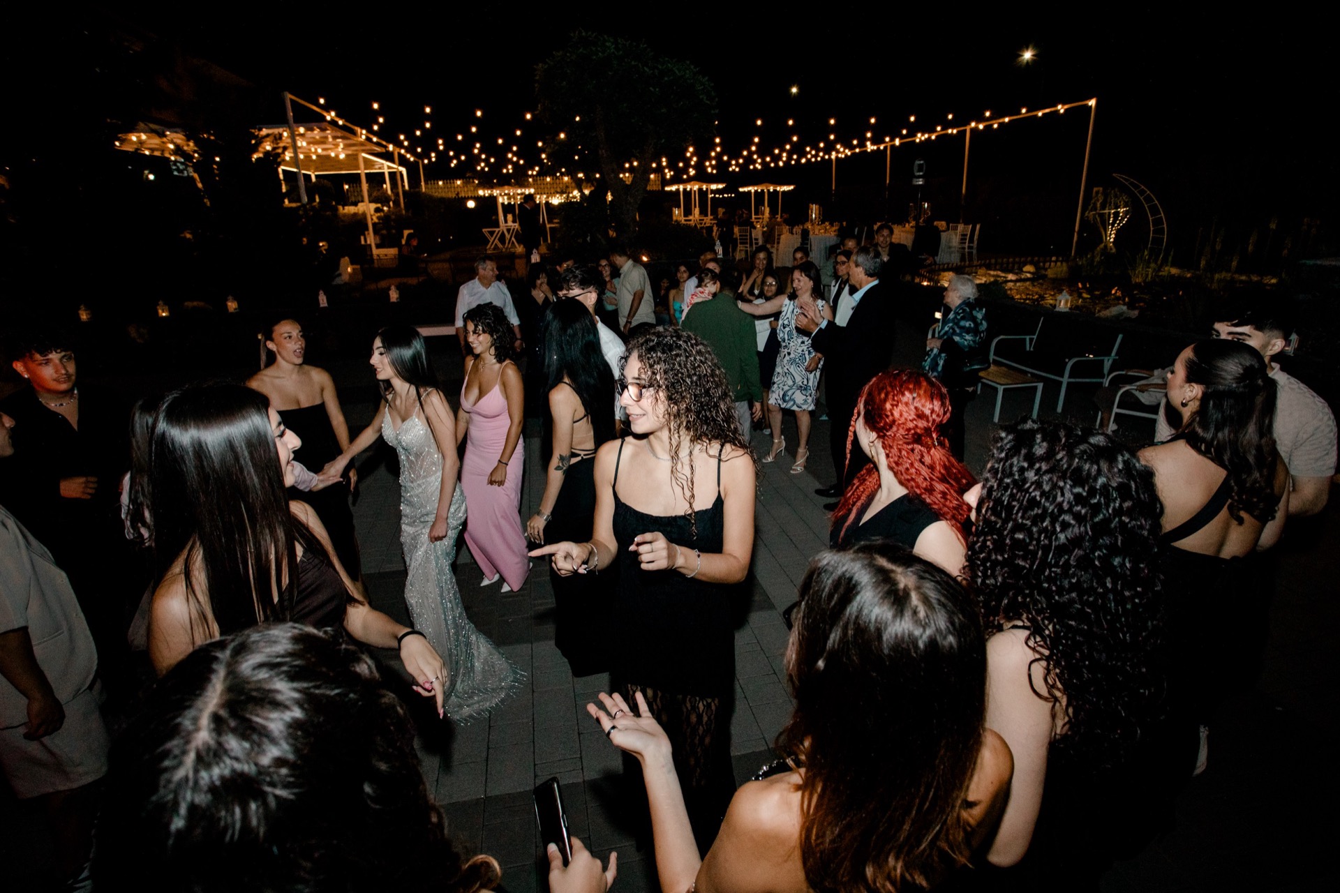 Pista da ballo all'aperto con ospiti che danzano di notte sotto una fila di luci sospese nel giardino. Sullo sfondo si intravedono tavoli apparecchiati, sedute bianche e un allestimento elegante da evento privato.