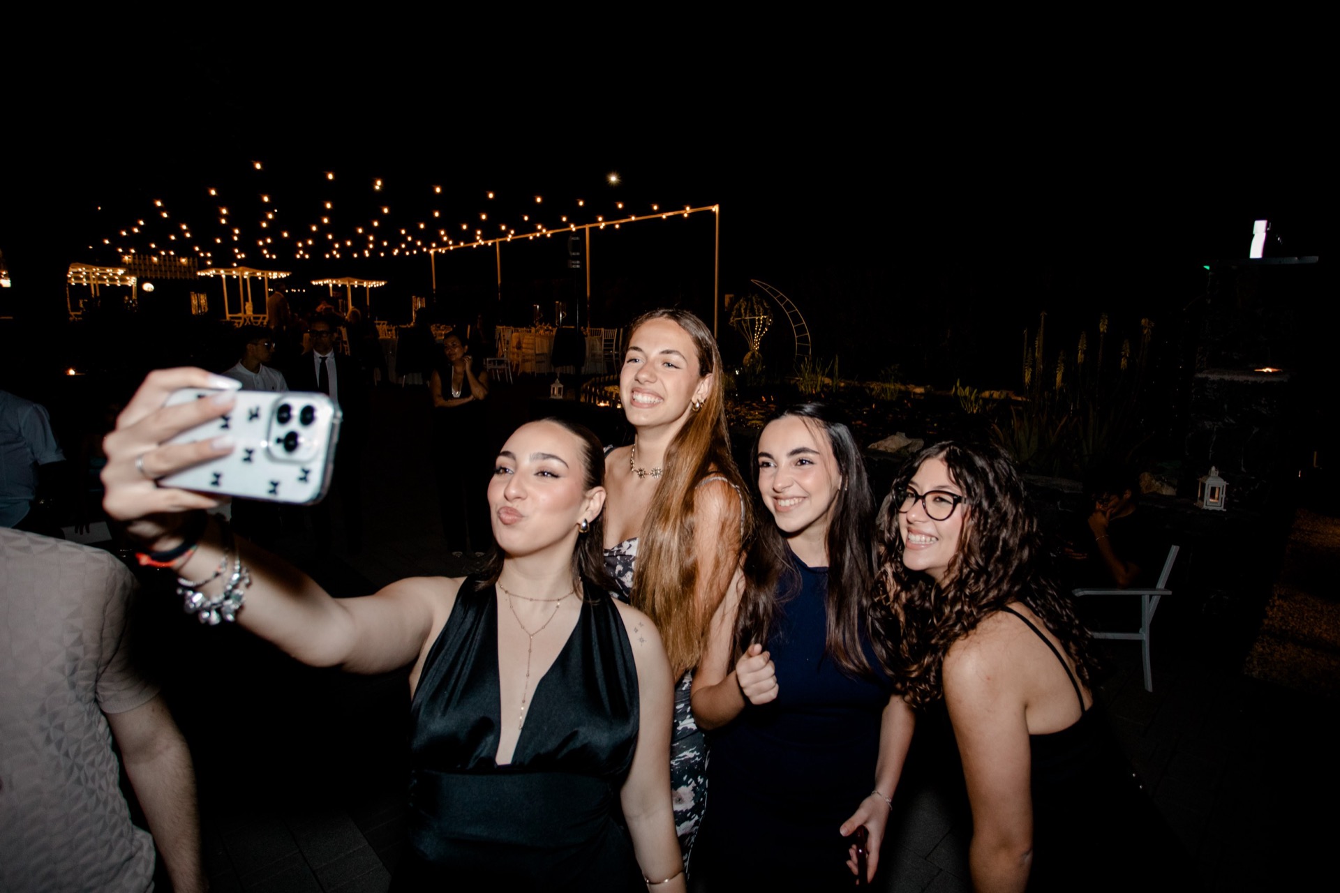 Gruppo di ospiti che si fa un selfie durante una festa serale all'aperto, con le luci del giardino e l'allestimento dell'evento sullo sfondo. L'immagine comunica convivialità e atmosfera informale.