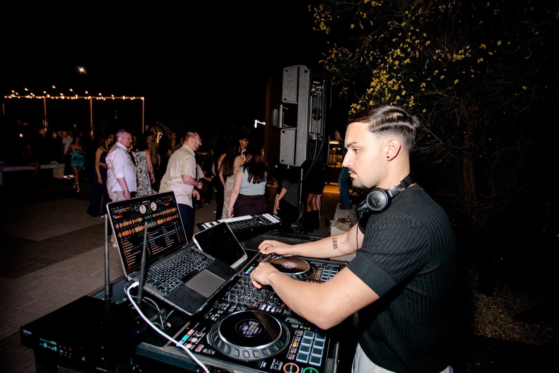 Postazione DJ durante una festa serale all'aperto, con console, laptop e casse professionali in primo piano. Sullo sfondo si vede la pista con ospiti in movimento e le luci calde sospese nel giardino.