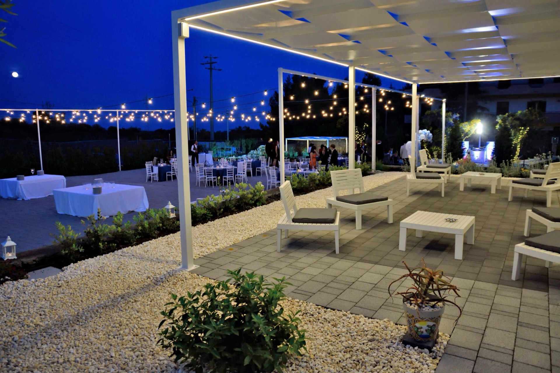 Area relax esterna con divanetti bianchi, tavolini bassi e pergolato moderno illuminato da luci calde. Sullo sfondo si vedono i tavoli dell'evento e il giardino allestito per la serata.