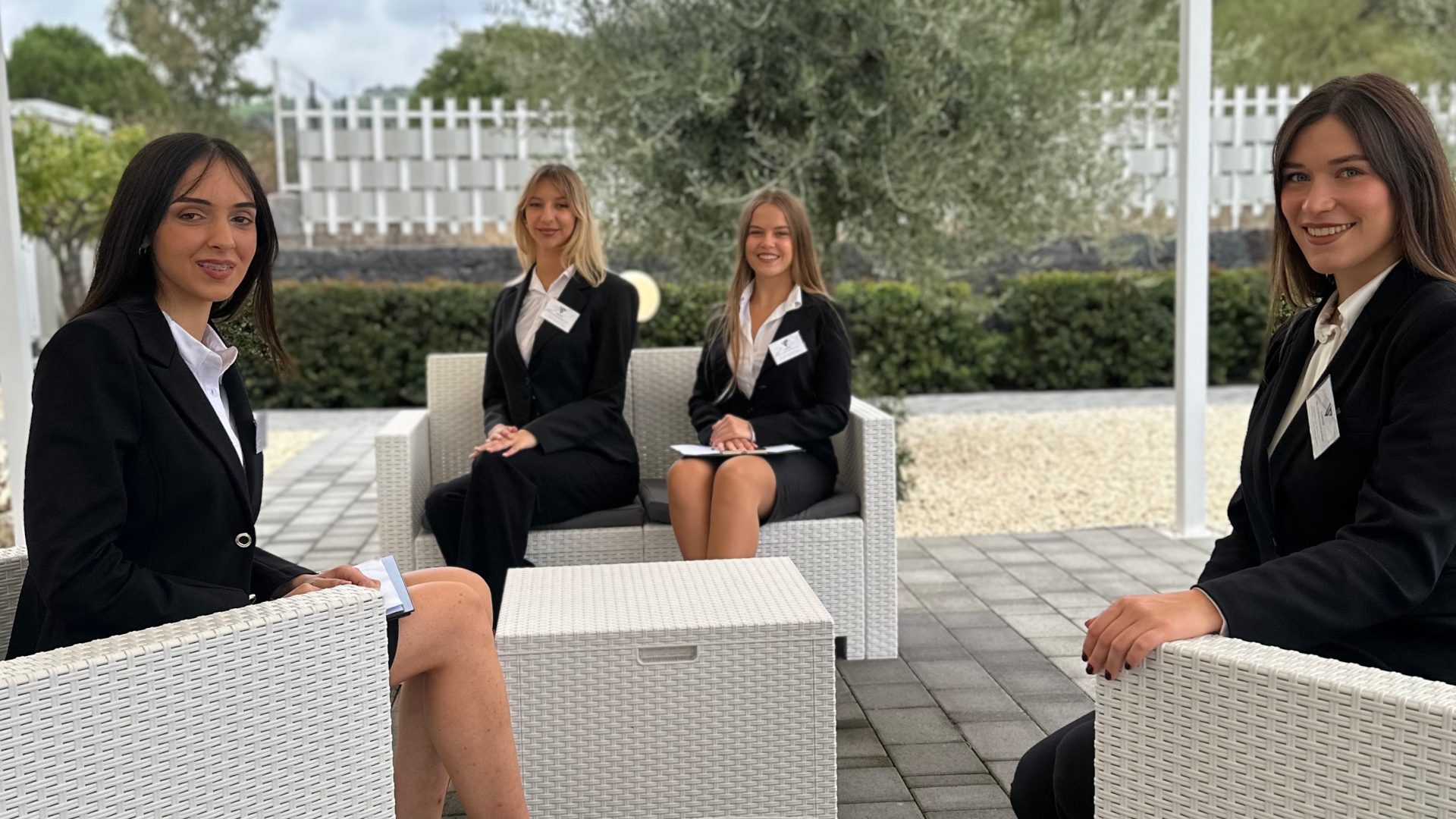 Angolo lounge esterno con quattro persone dello staff sedute su poltrone intrecciate e divanetto coordinato, tutte in divisa elegante. Sullo sfondo si vede il giardino e l'allestimento dell'area evento in pieno giorno.