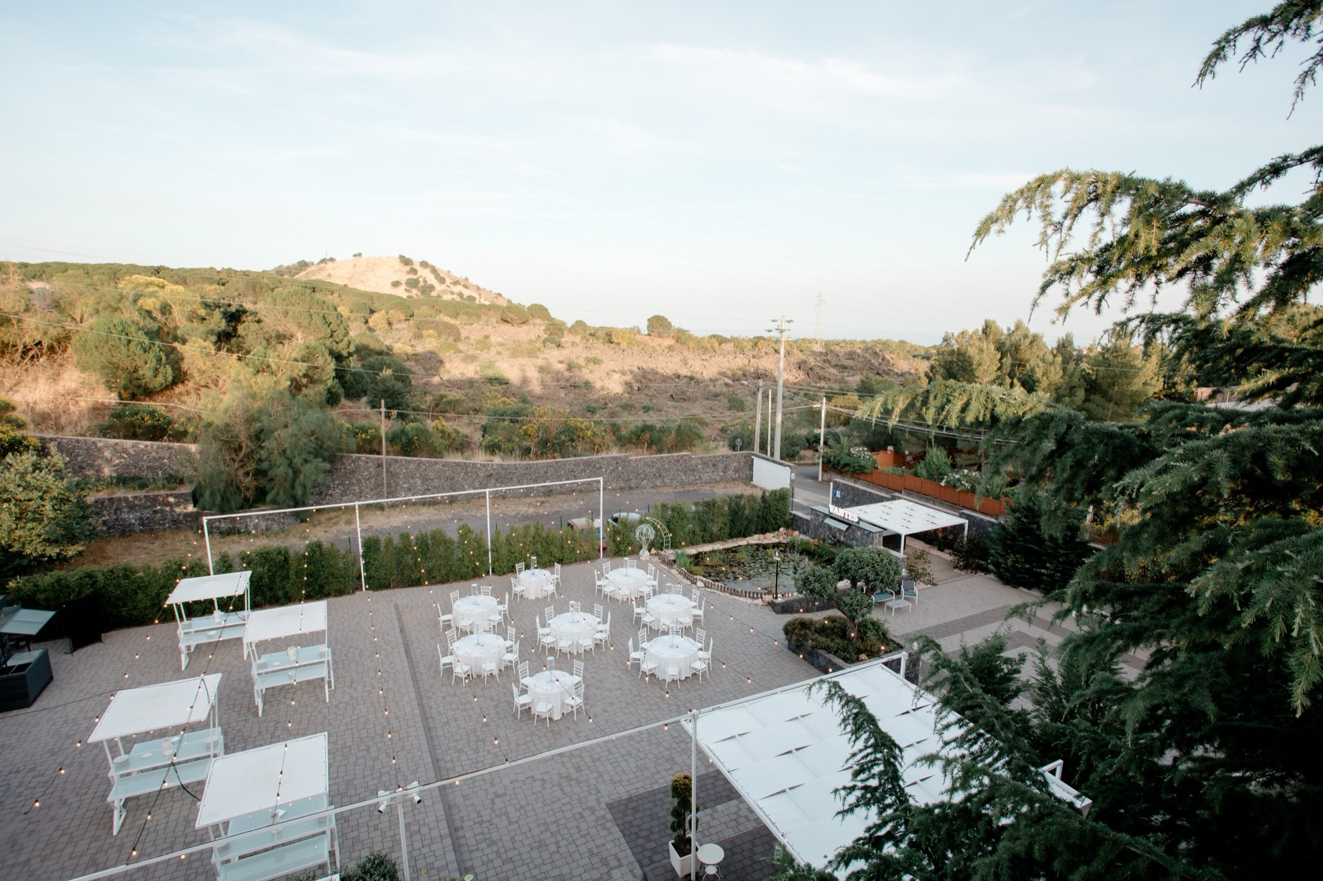 Panoramica diurna della location con tavoli rotondi, pergolati, aree buffet e vialetti curati distribuiti nel cortile. Sullo sfondo si vede il paesaggio collinare che circonda lo spazio evento.