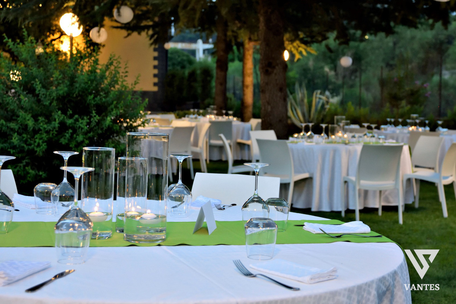 Cena all'aperto con tavoli apparecchiati, tovagliato bianco, runner verde e calici trasparenti sotto alberi illuminati. Sullo sfondo si vedono altre tavolate e l'atmosfera rilassata di un ricevimento serale.