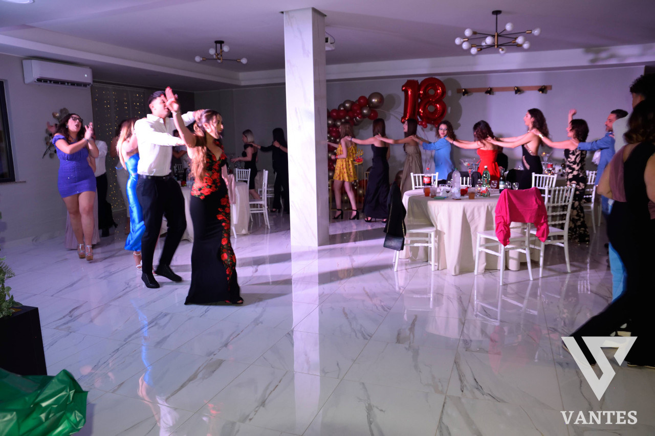 Festa dei 18 anni in sala interna con invitati che ballano in catena e tavoli apparecchiati sullo sfondo. Sul fondo si vedono palloncini rossi con il numero 18 e l'atmosfera è luminosa e movimentata.