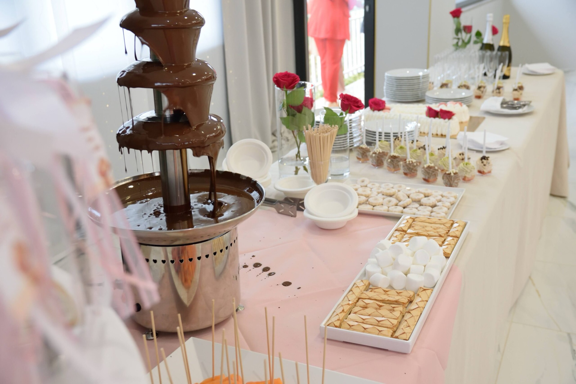 Fontana di cioccolato su tavolo buffet con marshmallow, biscottini e piccoli dolcetti disposti in vassoi ordinati. Accanto alla fontana ci sono piattini, bicchieri e decorazioni floreali per la postazione dessert.