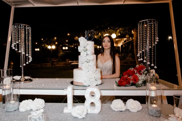 Ragazza in abito elegante accanto a un tavolo torta all'aperto di sera, sorridente mentre tiene un bouquet di rose rosse. Al centro si vede una torta bianca a più piani con decorazioni floreali e topper 18, affiancata da composizioni di rose bianche, candele e dettagli luminosi sotto una struttura coperta.