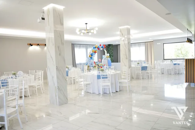 Sala luminosa con tavoli rotondi apparecchiati con tovaglie bianche, sedie bianche e runner azzurri, ripresa in una prospettiva ampia e ordinata. Al centro della sala c'è un arco di palloncini colorati, con tavolo decorativo sul fondo, grandi finestre, pilastri chiari e pavimento lucido che riflette la luce.