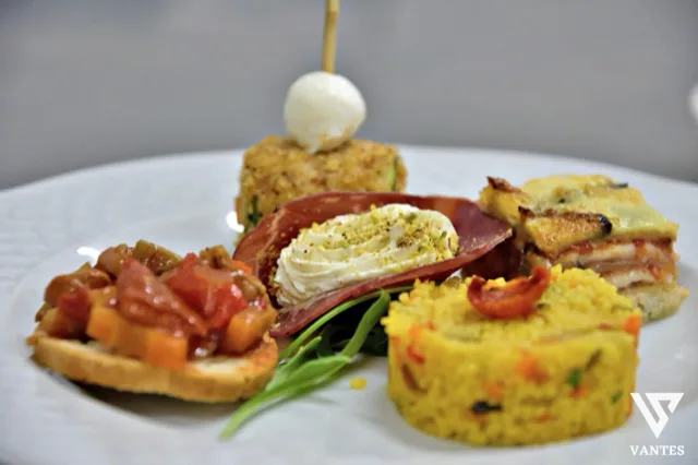 Primo piano di un piatto bianco con quattro piccoli assaggi salati disposti in modo armonioso: una tartina con verdure, una crema decorata con pistacchio, un cubetto di preparazione al forno e una porzione gialla impiattata con cura. Il piatto è guarnito con foglie verdi e mostra un dettaglio ravvicinato della proposta gastronomica.