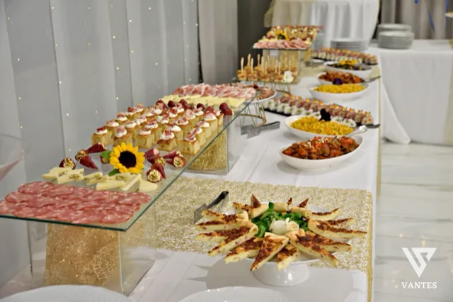 Lungo buffet salato allestito su più livelli con salumi, formaggi, tartine, spiedini, piccole portate calde e vassoi di antipasti disposti in file ordinate. Sul fronte ci sono piatti decorativi con assaggi a spicchi, mentre sullo sfondo si vedono piatti impilati e una tovaglia bianca con dettagli dorati.