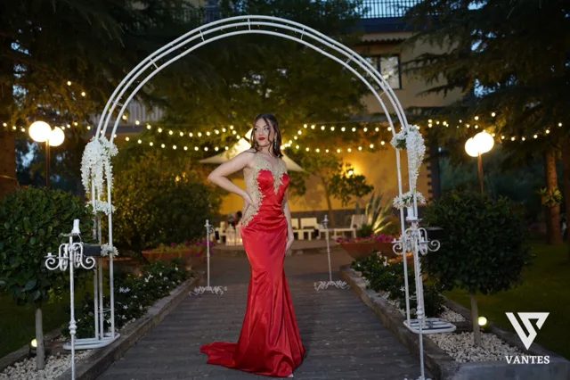 Ragazza in abito lungo rosso con dettagli dorati che posa al centro di un viale esterno decorato con un grande arco bianco, candele e fiori ai lati. Sullo sfondo si vedono le luci calde del giardino serale, i cespugli curati e la facciata della villa in penombra.