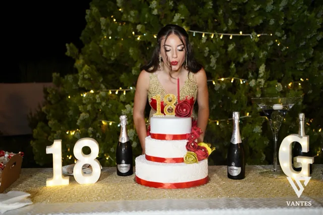 Tavolo torta all'aperto di notte con festeggiata in abito rosso e oro che soffia sulle candeline davanti a una torta a tre piani bianca e rossa. Sul tavolo ci sono numeri luminosi 18, bottiglie di spumante, un bicchiere cocktail a coppa e decorazioni floreali rosse e gialle su sfondo di siepe illuminata.