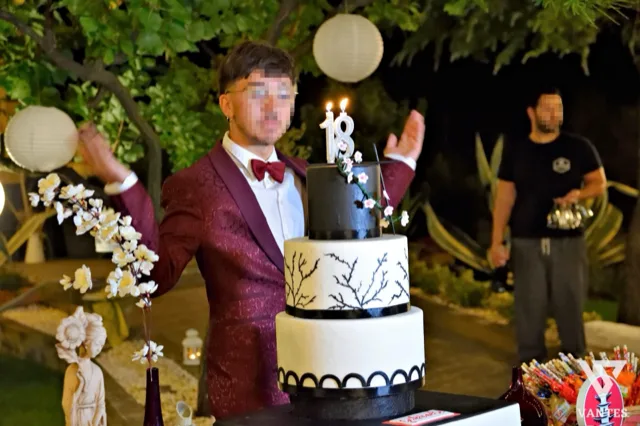 Ragazzo in giacca bordeaux accanto a una torta a tre piani bianca e nera con candeline a forma di 18, durante un festeggiamento serale in giardino. Sullo sfondo si vedono lanterne di carta appese agli alberi, piante ornamentali e un addetto di sala in un'atmosfera raccolta da taglio torta.