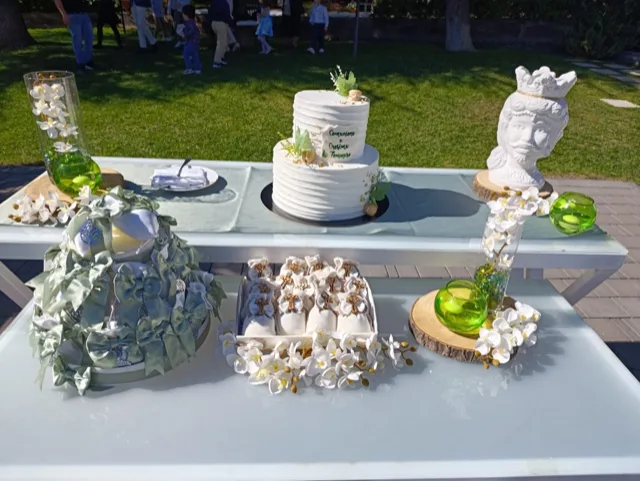 Tavolo torta per Comunione e Cresima con torta bianca a due piani, bomboniere in primo piano, vasi verdi con fiori bianchi e una testa di moro decorativa sul lato. L'allestimento è disposto all'aperto davanti al prato, con dettagli chiari e raffinati dedicati alla cerimonia.
