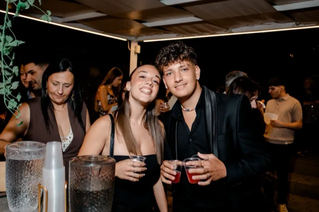 Area aperitivi all'ingresso con due giovani in primo piano che tengono bicchierini e sorridono verso la camera, mentre altri ospiti sostano dietro al banco beverage. Sul tavolo si vedono contenitori per drink, bicchieri e accessori da servizio sotto una copertura illuminata di sera.