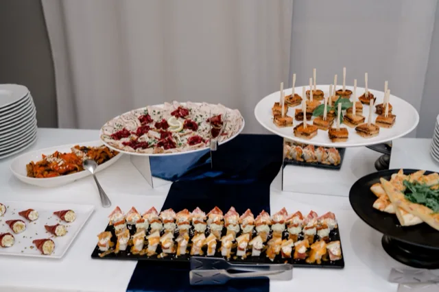 Tavolo buffet centrale con numerose portate fredde e calde disposte su alzate di vetro, piatti bianchi e vassoi neri. Si vedono finger food, salumi, formaggi, mini tramezzini e antipasti monoporzione in una presentazione ricca e ordinata da catering.