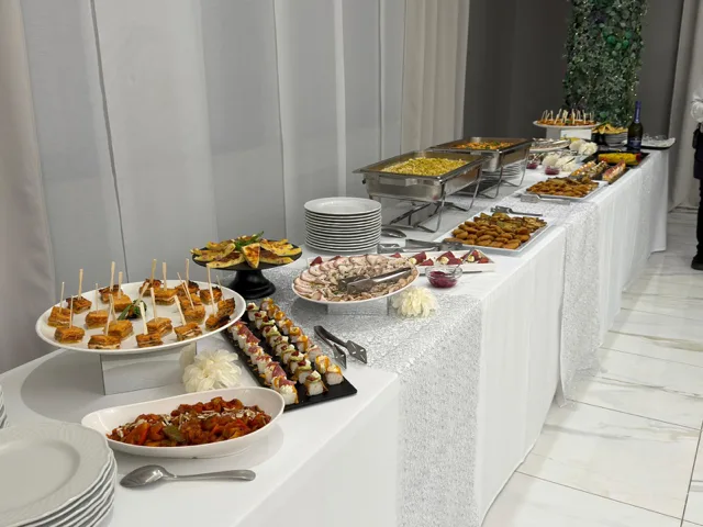 Buffet caldo in sala con vasche inox di primi e contorni, piatti impilati ai lati e vassoi di fritti e antipasti disposti sul tavolo bianco. La scena mostra una postazione di servizio ordinata e luminosa, con toni neutri e allestimento minimale.