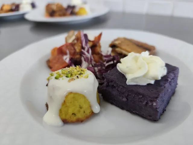 Primo piano di un piatto con piccoli assaggi gourmet, tra cui un tortino giallo con salsa bianca, una porzione viola con crema in superficie e altre preparazioni salate sullo sfondo. L'inquadratura ravvicinata mette in evidenza contrasti di colore, consistenze e cura dell'impiattamento.