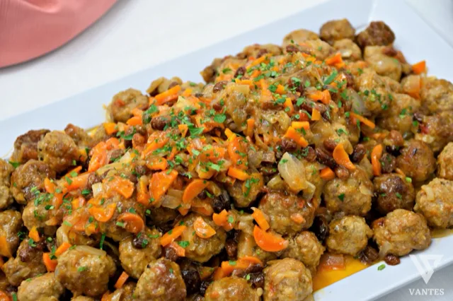 Grande vassoio di polpettine servite con un condimento ricco di cipolla, carote, uvetta e salsa lucida, ripreso in primo piano. La portata ha un aspetto generoso e casalingo, pensato per il servizio buffet di un secondo piatto caldo.