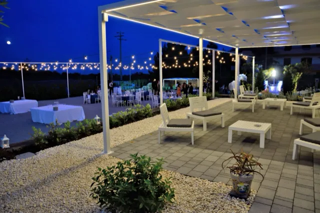 Area lounge esterna sotto un pergolato bianco, con sedute basse coordinate, tavolini e pavimentazione chiara circondata da ghiaia e verde ornamentale. Sullo sfondo si vedono i tavoli dell'evento e le catenarie luminose che accendono la scena serale.