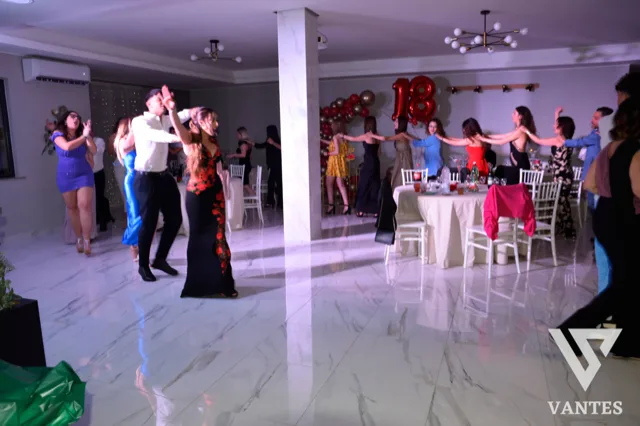 Festa dei 18 anni in sala interna con invitati che ballano in catena e tavoli apparecchiati sullo sfondo. Sul fondo si vedono palloncini rossi con il numero 18 e l'atmosfera è luminosa e movimentata.