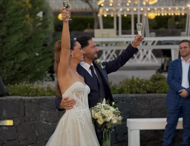 Sposi all'aperto durante il brindisi, con calici alzati e bouquet bianco tra le mani della sposa, ripresi davanti a muretto in pietra e luci decorative serali. L'immagine racconta un momento di festa elegante subito dopo la cerimonia o il ricevimento.