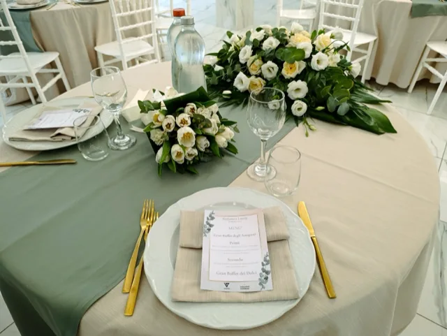 Dettaglio di mise en place matrimonio con piatti bianchi, posate dorate, menu personalizzato e runner verde salvia su tavolo tondo. Sul piano si vedono anche bouquet e composizione floreale bianca, in una sala luminosa dall'allestimento raffinato.