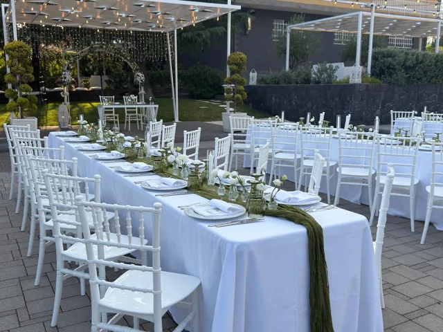 Tavolata imperiale e tavoli rotondi apparecchiati all'aperto per matrimonio, con sedie bianche, runner verde salvia e piccoli vasi floreali distribuiti lungo il centro tavola. Sullo sfondo si vede l'area del rito con arco floreale sotto le strutture illuminate.