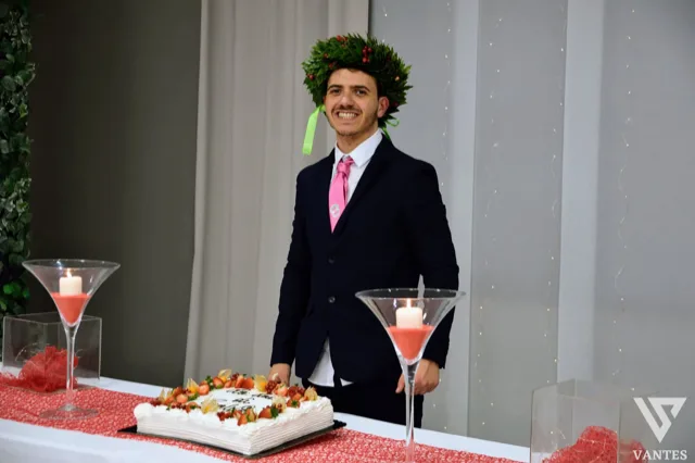 Ragazzo laureato in abito scuro con cravatta rosa e corona d'alloro, in posa sorridente dietro il tavolo torta durante il ricevimento. In primo piano compaiono una torta rettangolare decorata con fragole, due grandi candelieri con candele accese e dettagli rossi coordinati con l'allestimento.