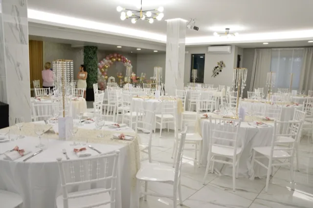Sala interna apparecchiata per cerimonia con tavoli rotondi bianchi, sedie chiavari, runner chiari e alti centrotavola pendenti. Sullo sfondo si vede un arco di palloncini nei toni del rosa e dell'oro con tavolo decorativo coordinato.
