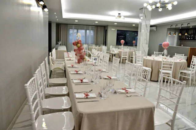 Sala apparecchiata per battesimo con una lunga tavolata centrale, tavoli rotondi sullo sfondo, sedie bianche e dettagli rosa nei nastri dei tovaglioli e nelle decorazioni. L'ambiente è luminoso e ordinato, con un allestimento delicato e coerente.