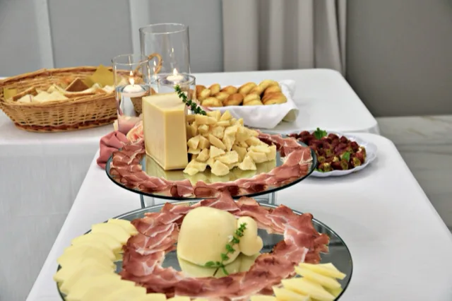 Angolo buffet con salumi e formaggi disposti su alzate a specchio, affiancati da pane, grissini e piccoli assaggi sullo sfondo. L'allestimento unisce presentazione decorativa e varietà di prodotti per il momento aperitivo o buffet.